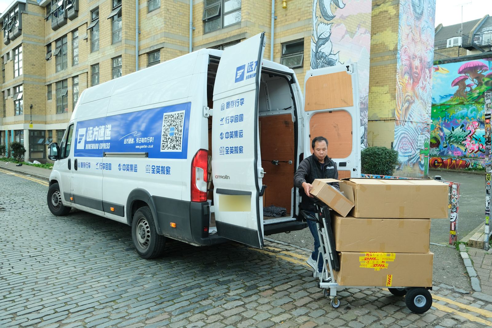 A Homeward Express driver loading boxes on a hand truck beside the company's white van, back doors open, on a cobbled London street.